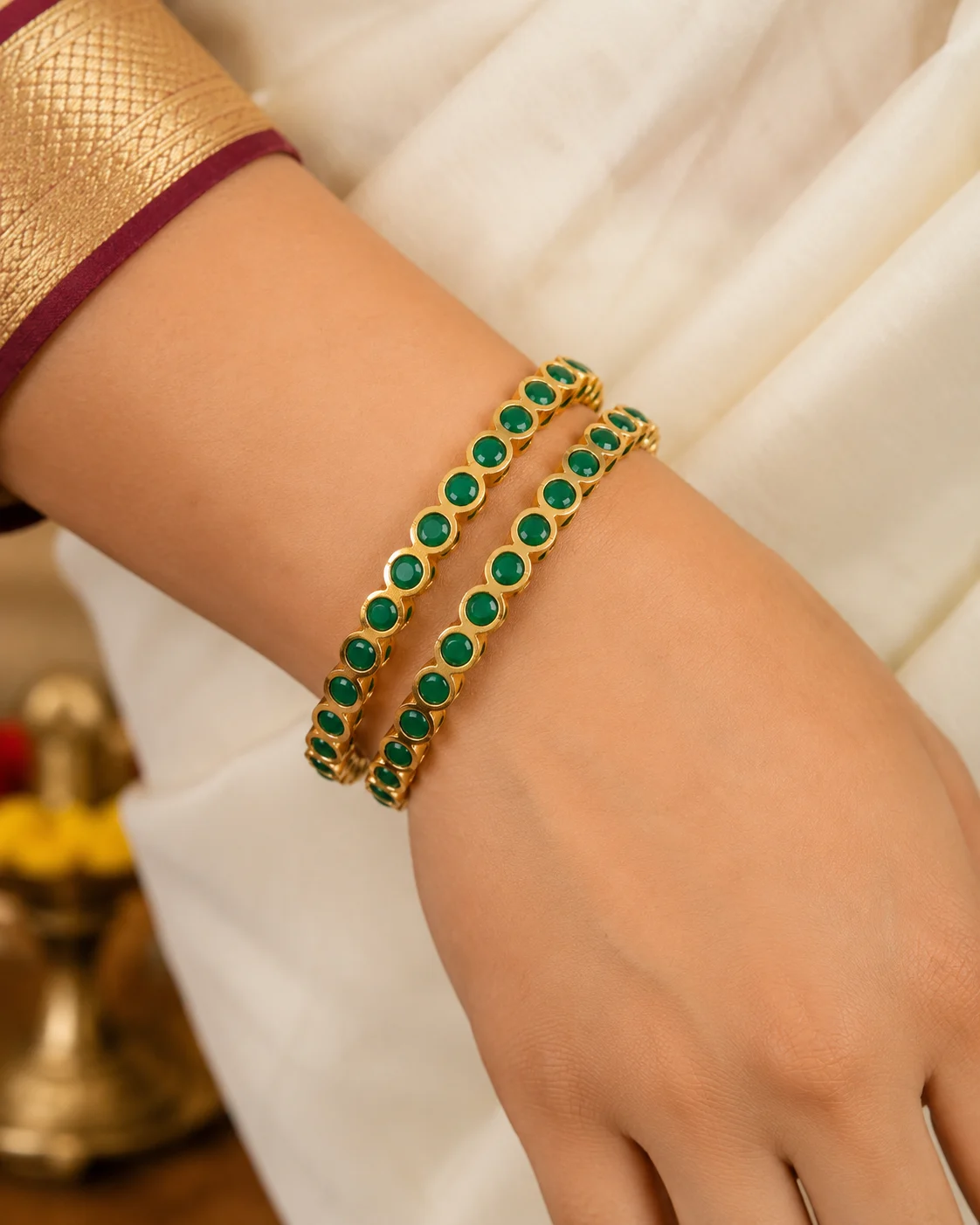 Pair of gold-tone bangles with round green accents worn on a wrist with a cream sari
