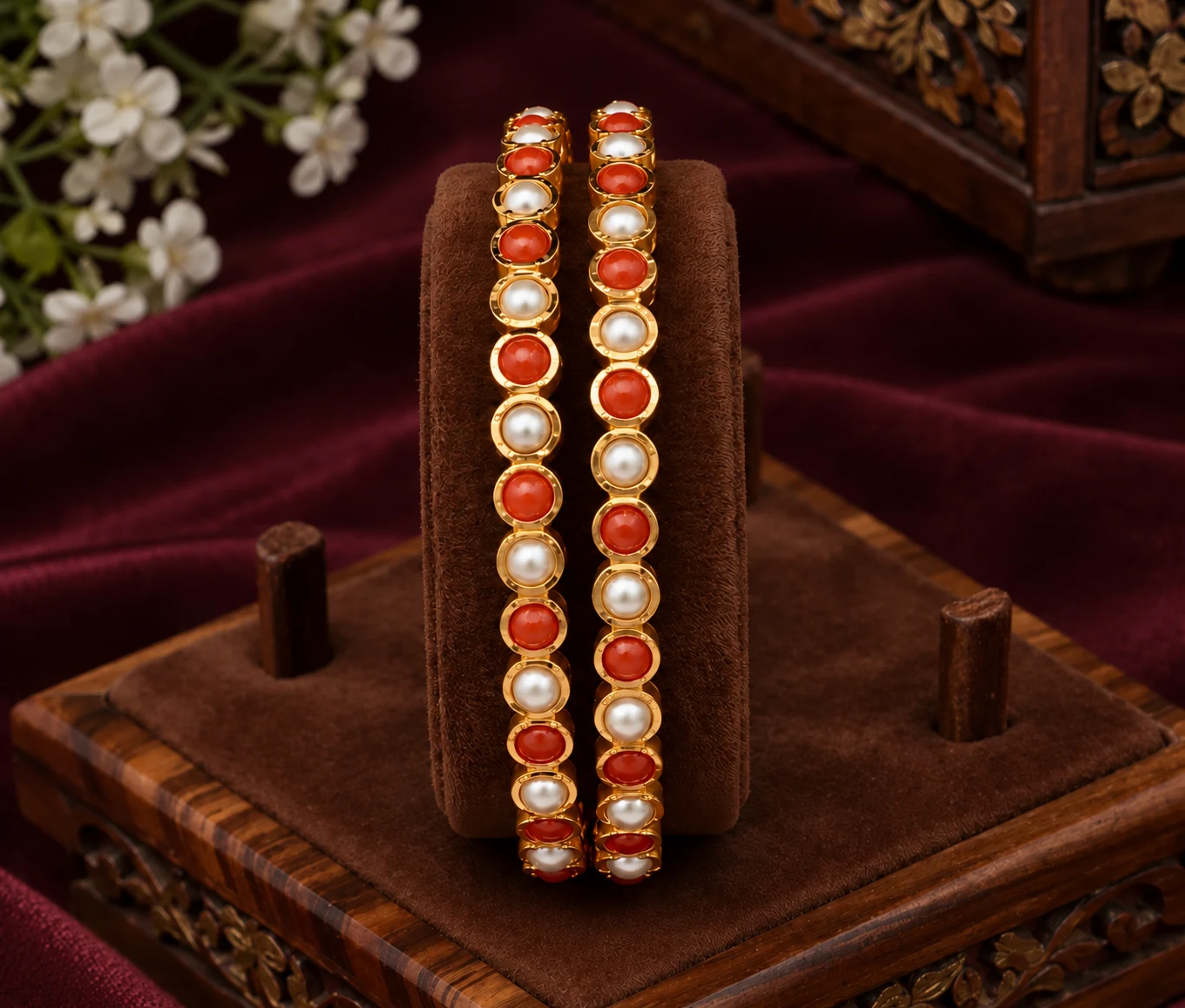 Pair of gold-tone bangles with alternating pearl-like and coral-orange round accents on a brown display stand